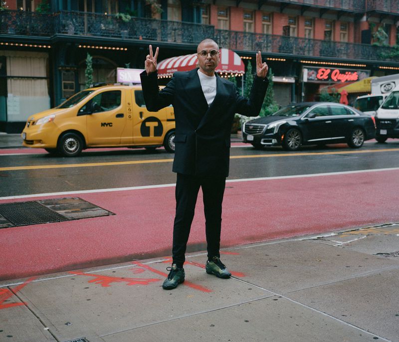 Man in a black suit jacket with peace signs on a red-marked crosswalk in a city street with yellow cabs and dark cars.