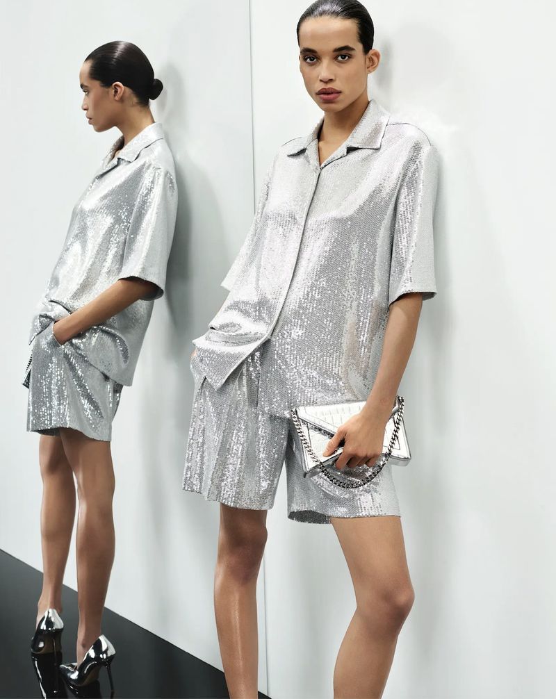 A woman in a shimmering silver sequined short set and heels poses against a reflective wall in a studio, holding a silver clutch.