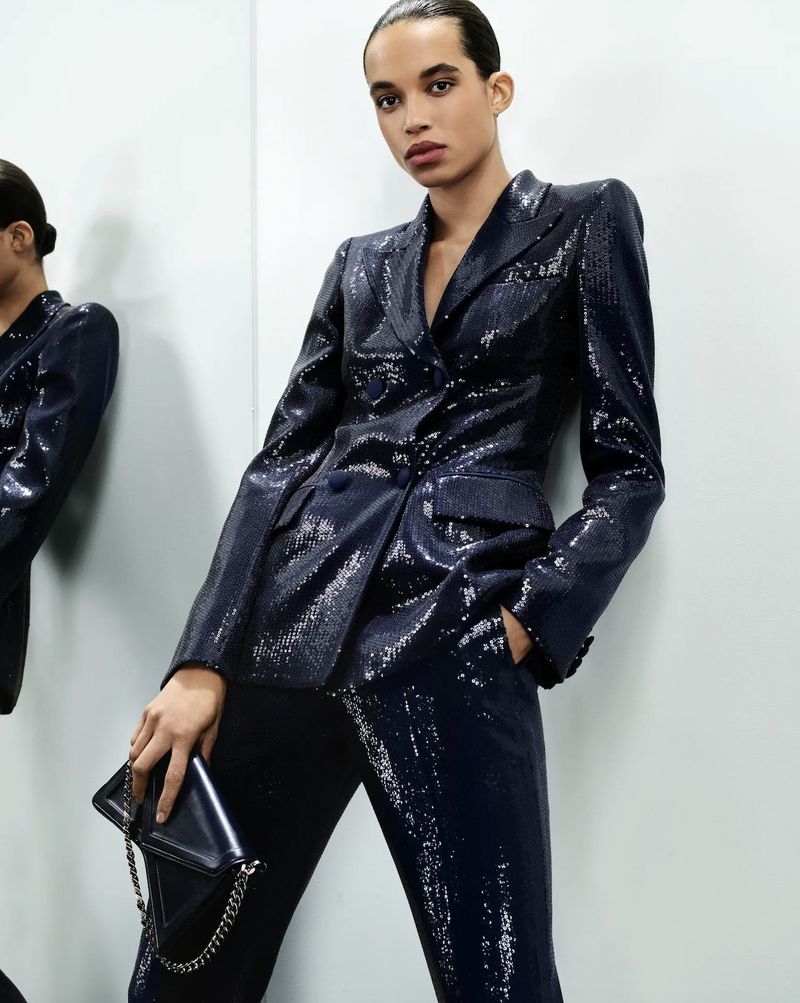 Fashion shoot of a woman in a dark, shiny double-breasted suit, holding a dark clutch, against a neutral wall with a partial mirror.