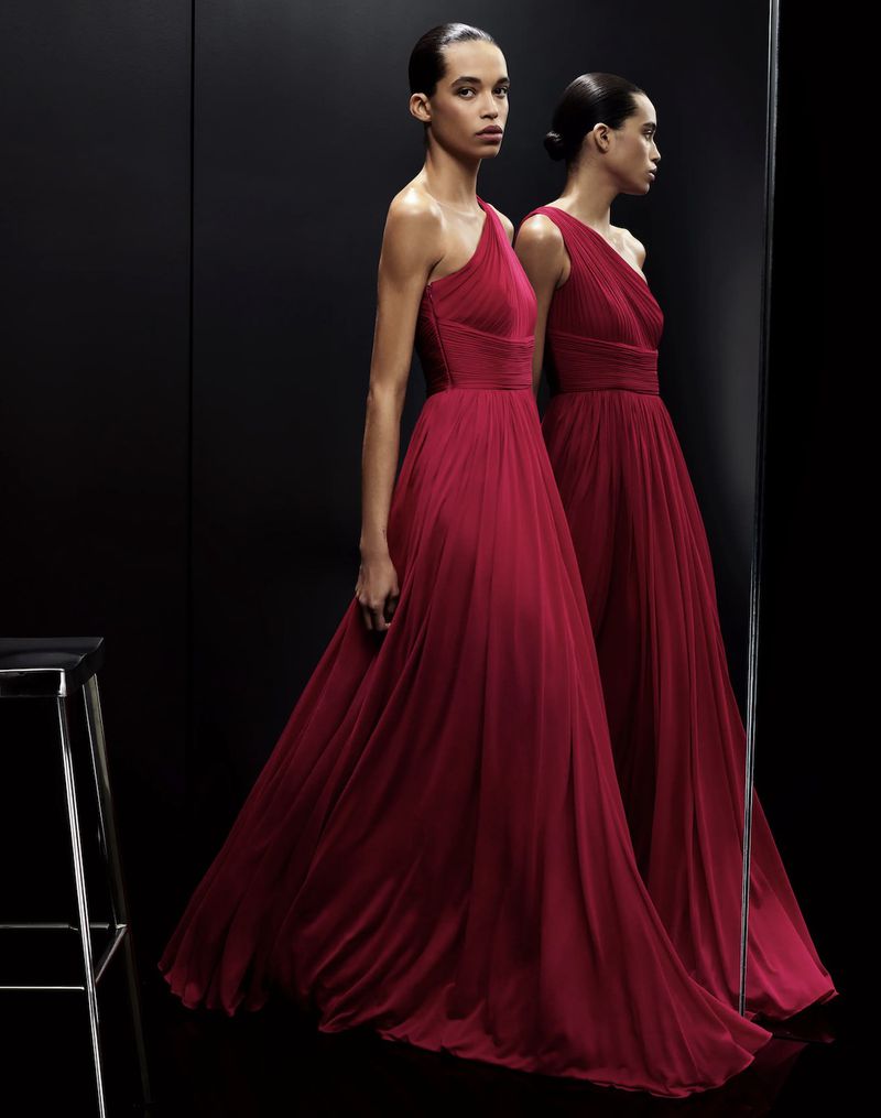 Two women wearing red formal dresses are reflected in a mirror in a dimly lit indoor studio setting. Fashion and portrait photography.
