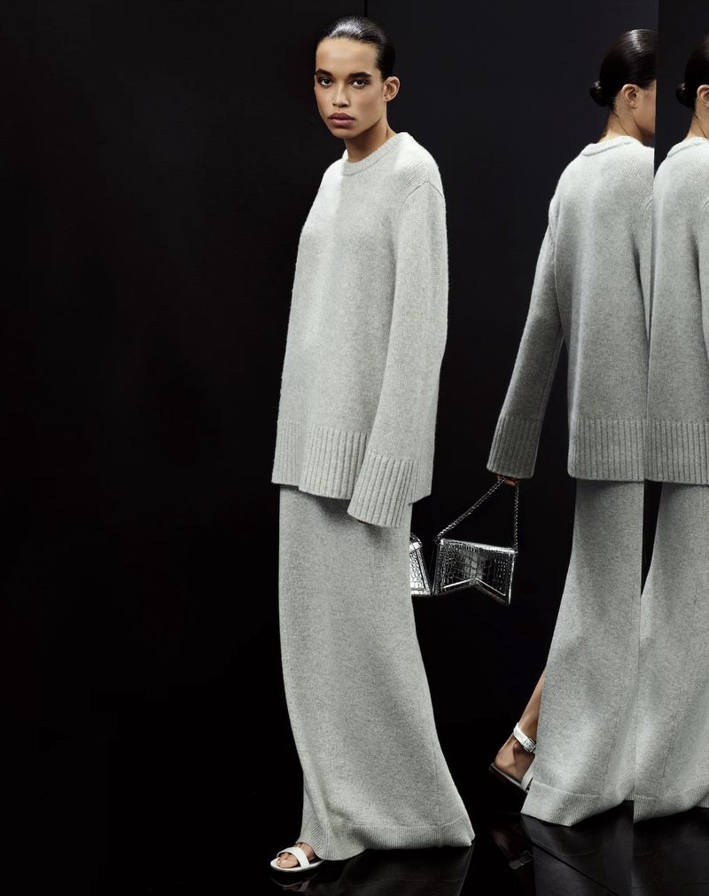 Woman in grey knitwear ensemble holds a silver purse, posed in a dark studio with multiple reflections. Fashion and minimalism.