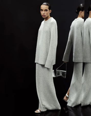 Woman in grey knitwear ensemble holds a silver purse, posed in a dark studio with multiple reflections. Fashion and minimalism.