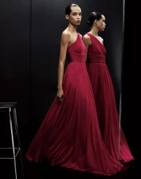 Model Lara Menezes wearing a pleated red one-shoulder evening gown in front of a black studio backdrop with reflection