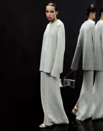 Model wearing a grey knit sweater and matching long skirt holding a silver handbag in a dark mirrored setting