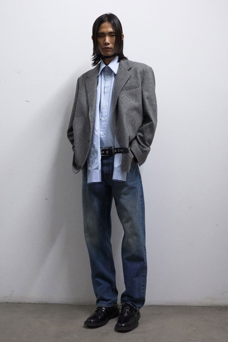 Asian man posing in a light blue dress shirt, gray tweed blazer, faded blue jeans, black belt, and black dress shoes in a studio.