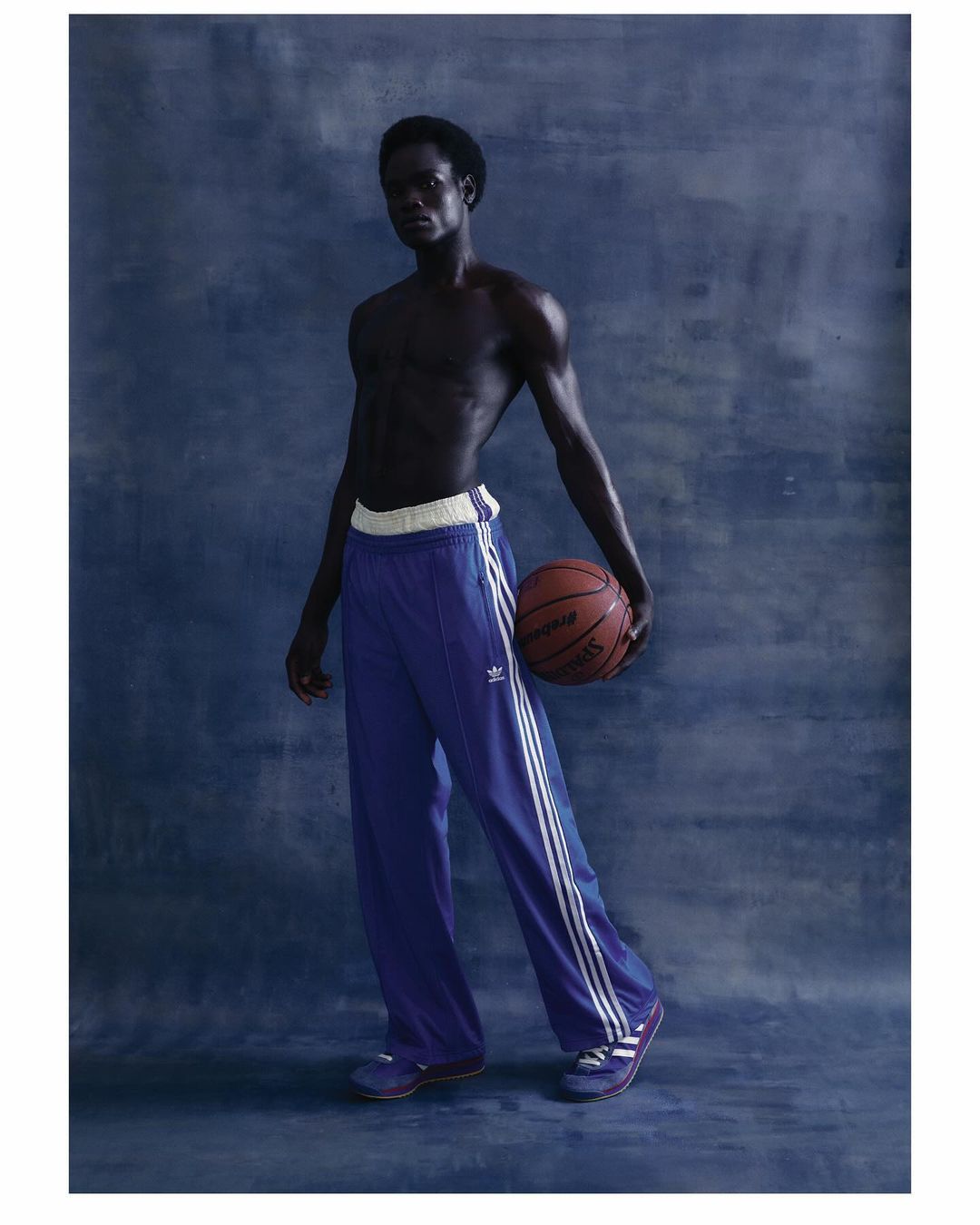 Athletic man in blue Adidas track pants holds a basketball in a studio setting with a textured blue backdrop, captured in a posed shot.