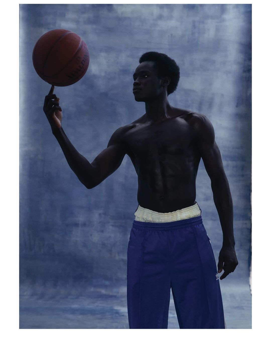 Athletic African American man balances a basketball on his finger against a cloudy blue backdrop in a studio portrait.