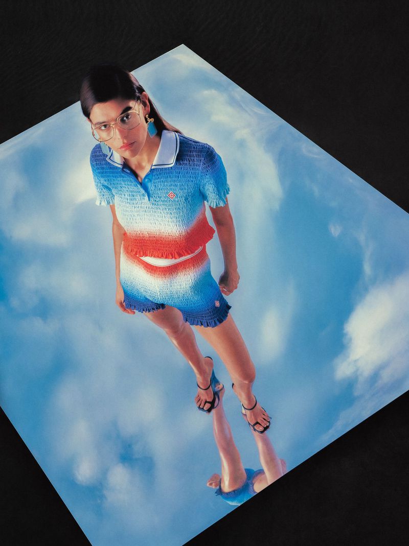 Woman in a red, white, and blue shorts outfit, wearing glasses, posing in front of a sky background reflecting on a glossy surface.