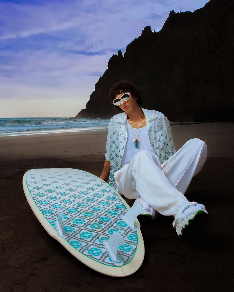 A male model poses on a beach next to a surfboard wearing sunglasses, a patterned shirt, white pants, and sneakers, with a mountain backdrop.