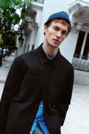 A young adult man wears a dark overshirt and blue beanie against an urban backdrop. Fashion shot in natural light.