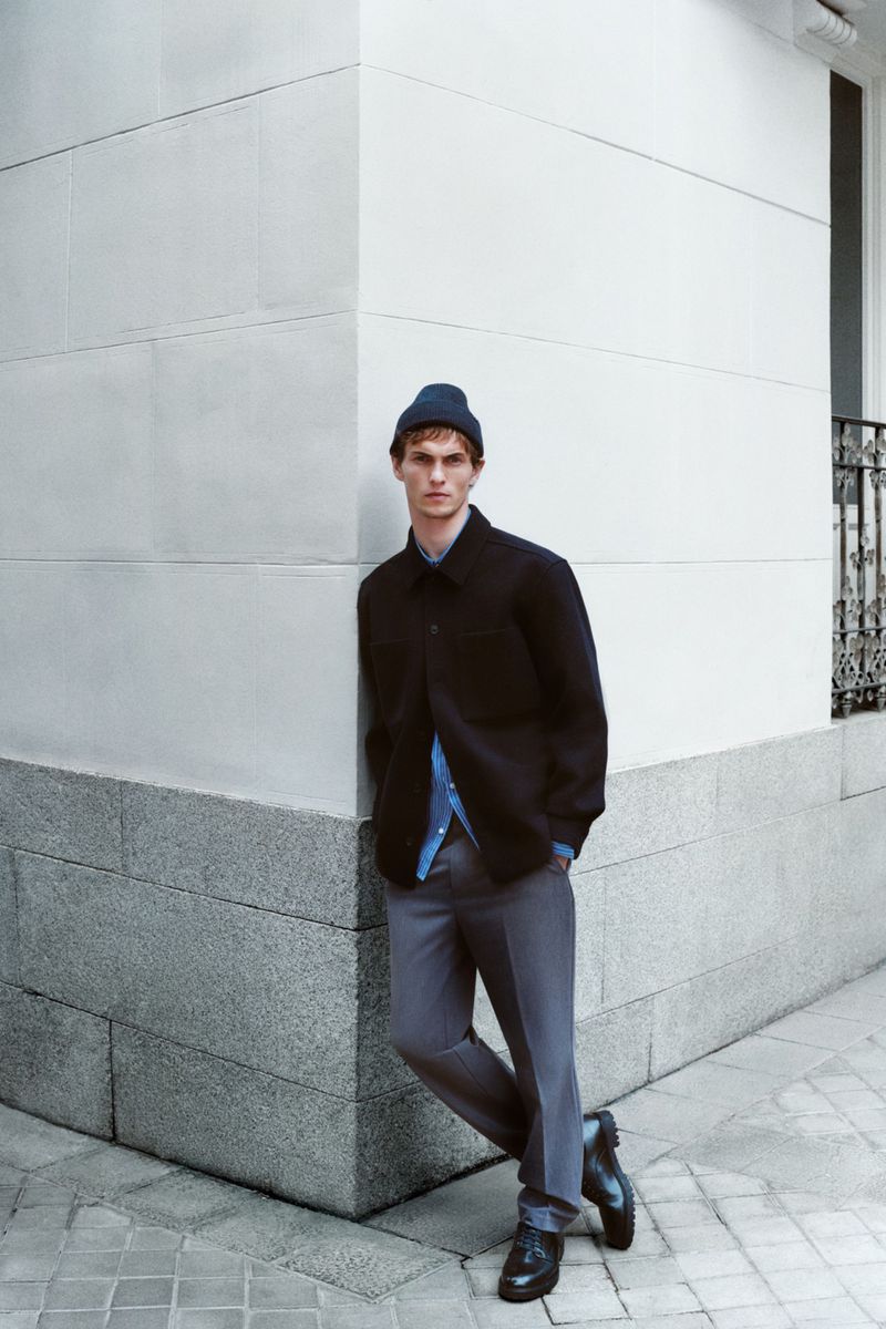 A young adult male model poses against a concrete wall wearing a navy beanie, a blue button-down shirt, a navy overshirt, grey trousers, and black boots.