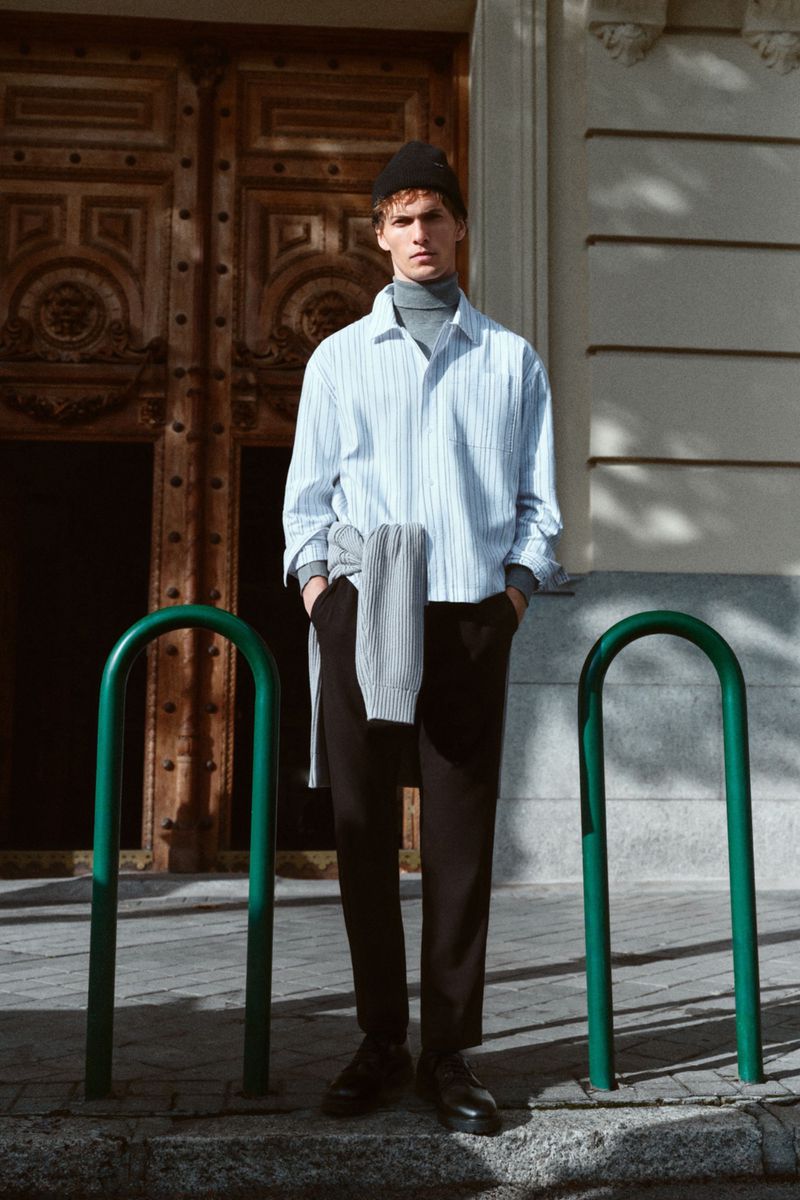 A young man in a striped shirt and beanie stands outdoors on a city street, with a sweater draped over his shoulder, posing confidently.