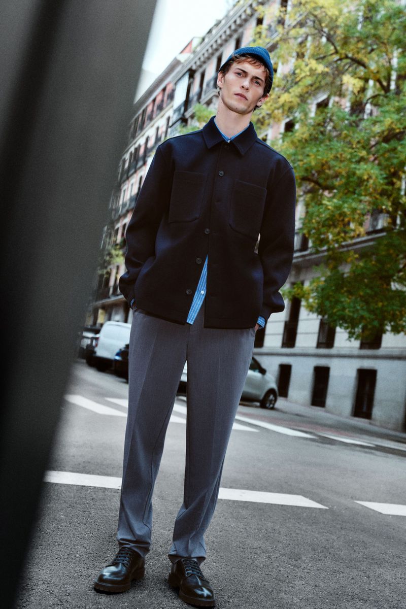 Stylish man in blue beanie, navy coat, grey pants, and black shoes stands on city street with cars and buildings in background.
