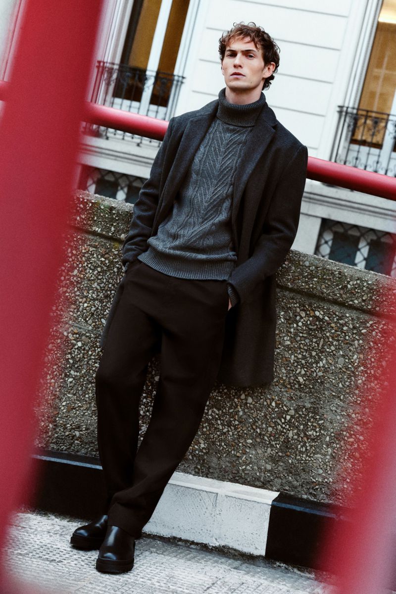 Model leaning against a stone wall wearing a charcoal coat, grey turtleneck sweater, and dark trousers.