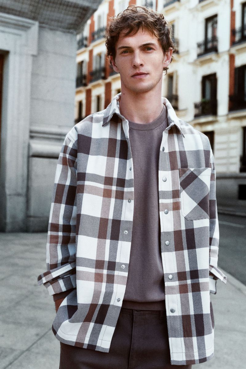 A young adult male poses outdoors wearing a brown and white plaid shirt over a brown t-shirt, standing on a sidewalk in an urban setting.