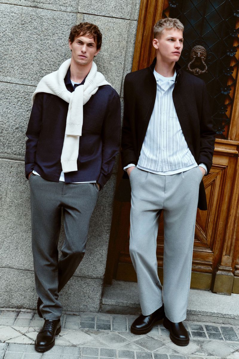 Two male fashion models in smart casual outfits pose outdoors. One wears a navy shirt and grey pants, the other a light shirt and blazer.