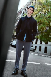A young male model in a dark shirt and grey trousers standing on a city street for the Lefties Timeless Wardrobe project.