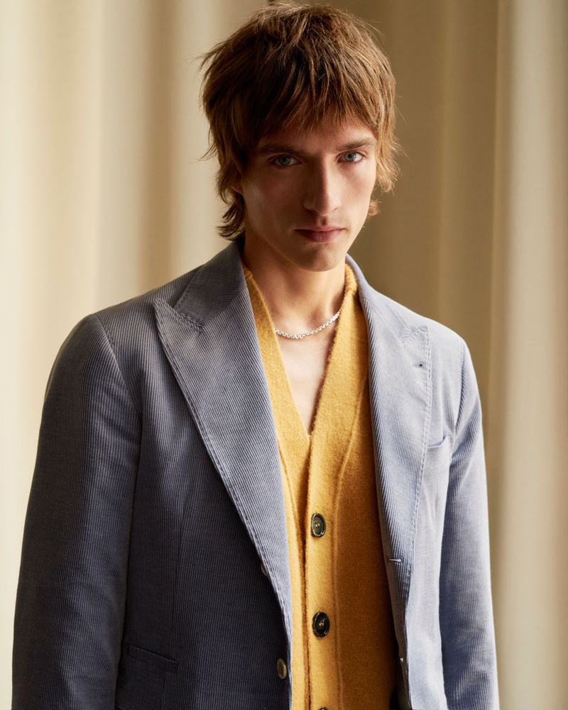 Portrait of a young adult male in a blue blazer and yellow cardigan, posing indoors against a light tan backdrop, with a neutral facial expression.