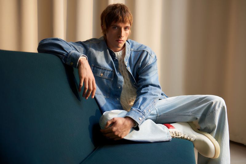 A male model in a denim shirt and wide-leg jeans poses on a blue couch in a fashion shoot. Cream-colored backdrop and warm lighting.