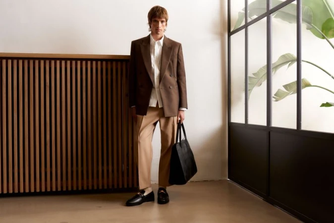 Adult male in blazer and khaki pants stands in an indoor setting with a black tote bag. Light neutral tones and a window view.