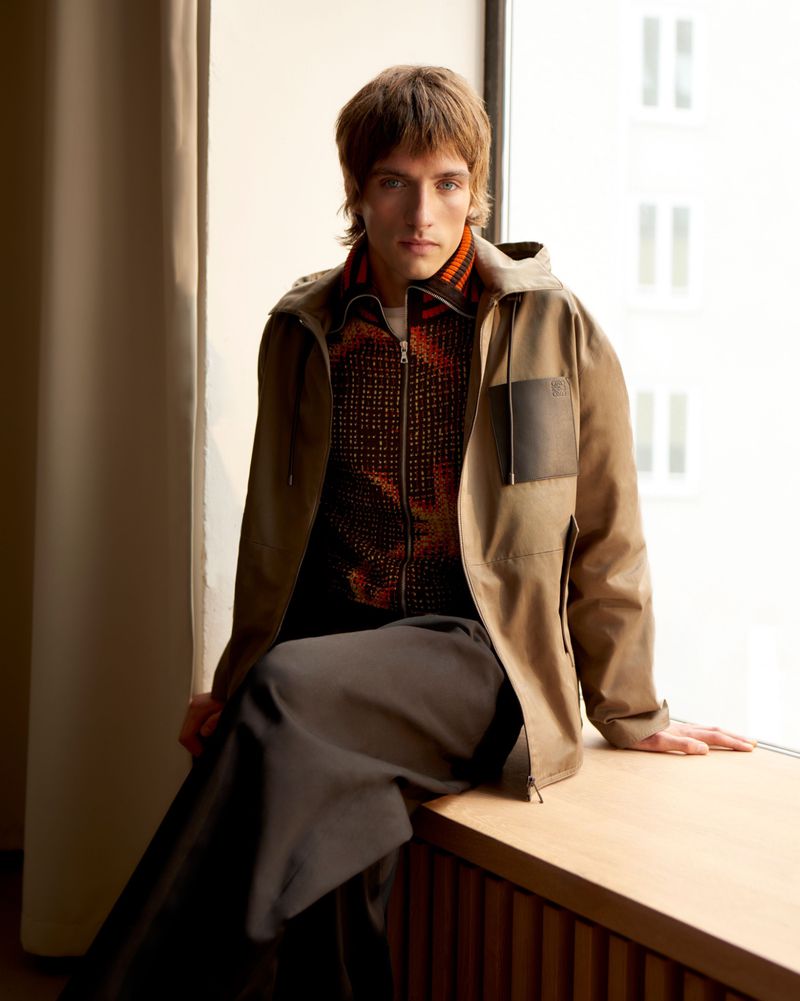 Stylish man in jacket sitting by the window. He is wearing a brown hooded jacket with dark trousers in an indoor setting.
