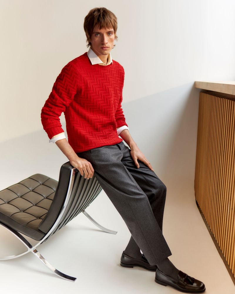 A stylish adult male model in a red sweater and gray trousers poses indoors on a black leather chair, showcasing men's fashion.