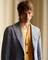 A young male model with shaggy brown hair wearing a blue blazer over a yellow cardigan against a neutral curtain background.