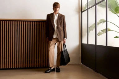 Model wearing a brown blazer, white shirt, and tan trousers while holding a black tote bag indoors by a slatted wooden wall.