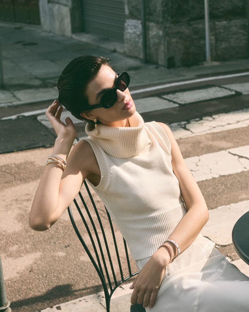 Woman in sleeveless top and sunglasses sitting outdoors in urban environment, accessorized with bracelets and earrings in daylight.