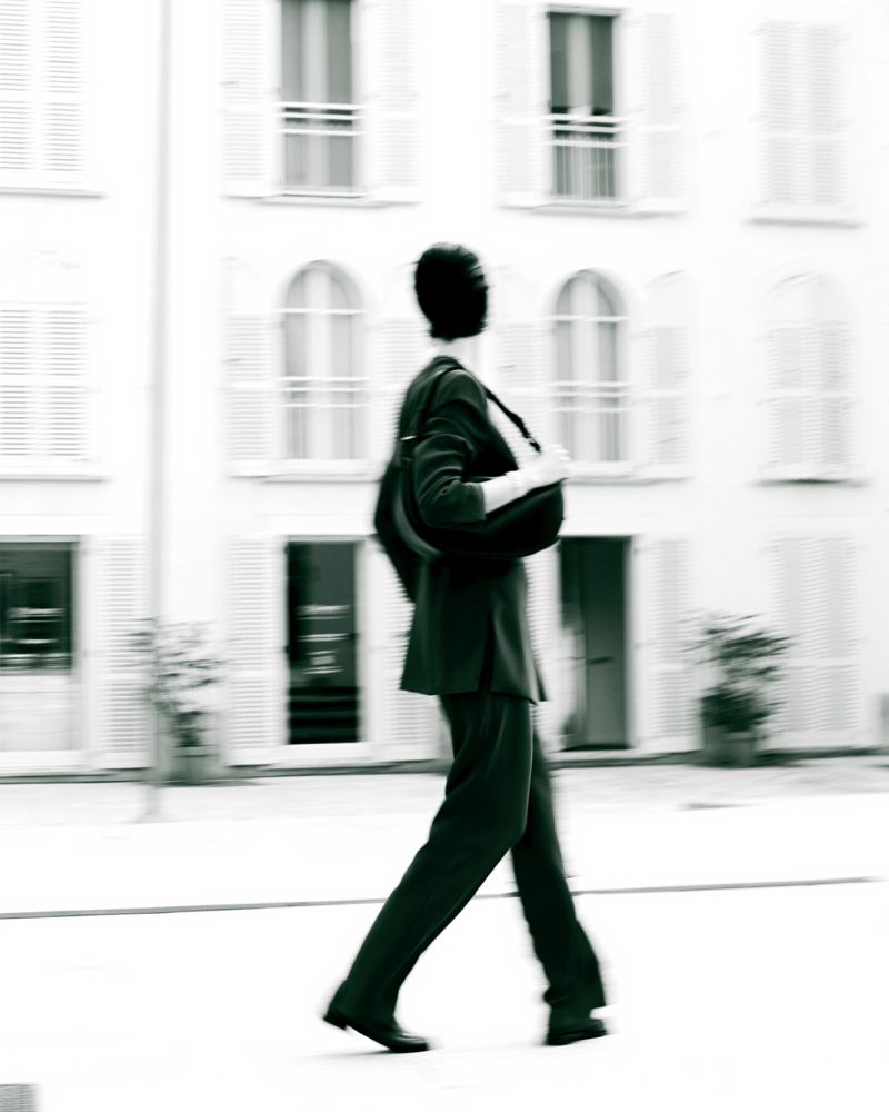 Model walking in motion blur wearing a dark jacket and trousers against a white building exterior