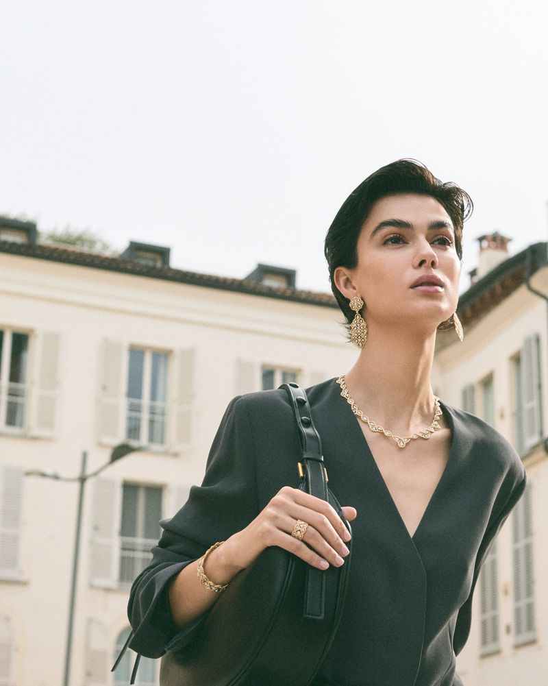 A woman with short dark hair poses outdoors in a dark grey jacket, wearing gold jewelry and carrying a black purse in daylight.