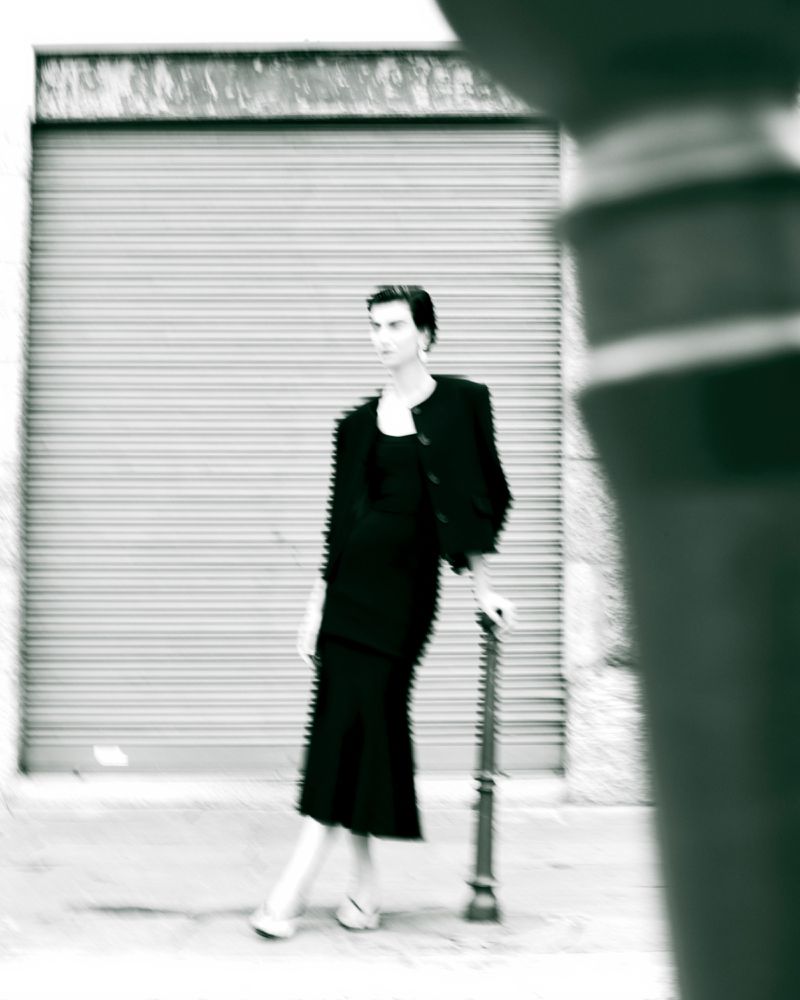 Fashionable adult female poses outdoors in a black suit dress with black cropped jacket, leaning against a lamp post with blurred motion.