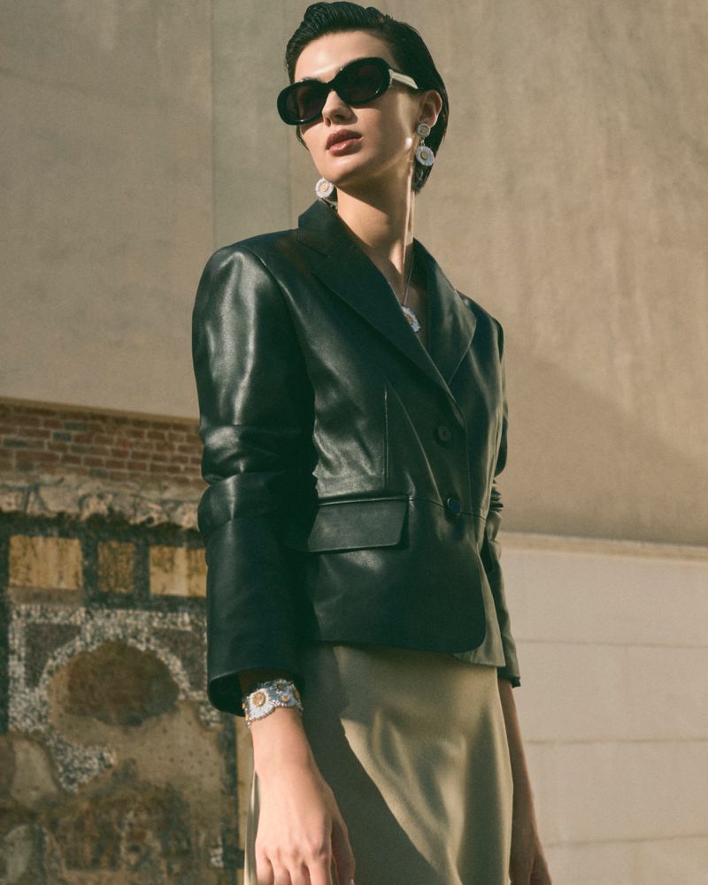 A woman with short hair and sunglasses poses outdoors wearing a black blazer, skirt, jewelry, and sunglasses in a fashion context.