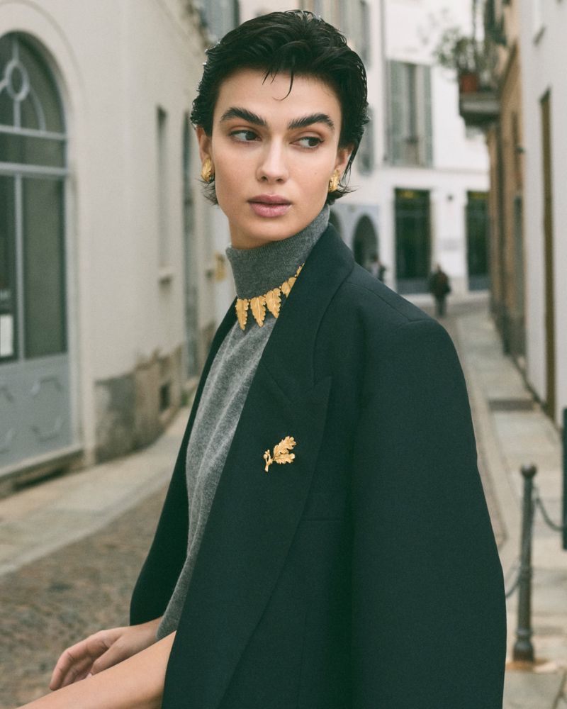 Brunette woman in a grey turtleneck, black blazer, and gold jewelry poses on a city street with buildings in the background.