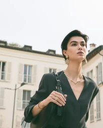 Model wearing gold jewellery and a black top carrying a black handbag outdoors against a traditional European building facade.