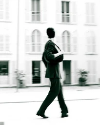 Model walking in motion blur wearing a dark jacket and trousers against a white building exterior