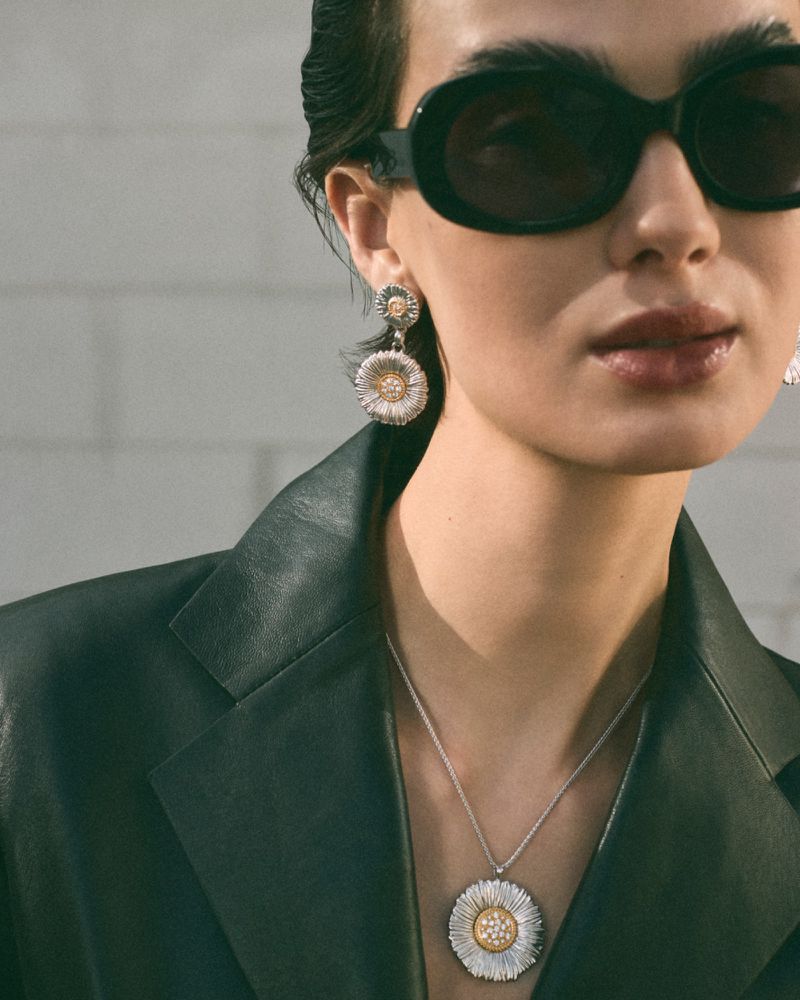Model wearing dark sunglasses, silver and gold flower pendant necklace, matching earrings, and a dark leather jacket against a light brick wall