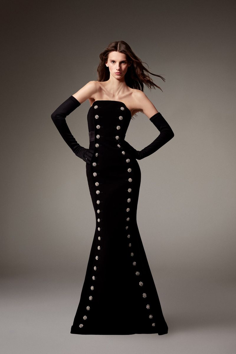 Model in strapless black gown with decorative silver buttons and long matching gloves against a grey studio backdrop.