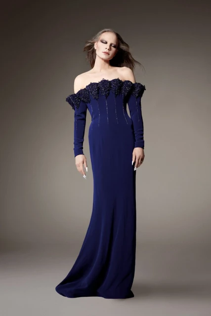 Elegant adult female with long hair, wearing a long-sleeved, off-the-shoulder blue gown in a full-length shot against a grey backdrop.