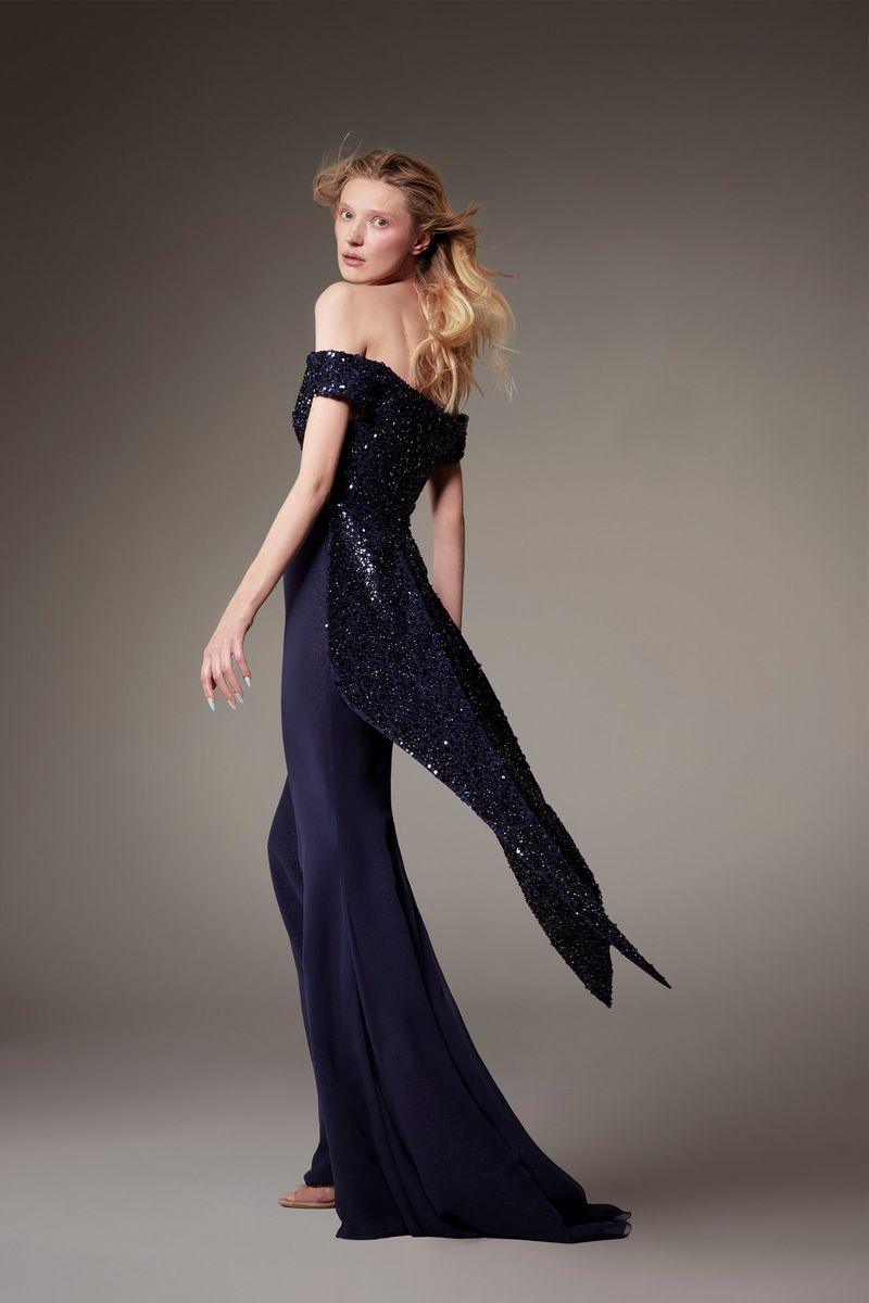 Blonde woman in an elegant off-the-shoulder navy blue gown poses in a studio. The gown has a sparkling sequin top and a long tail.