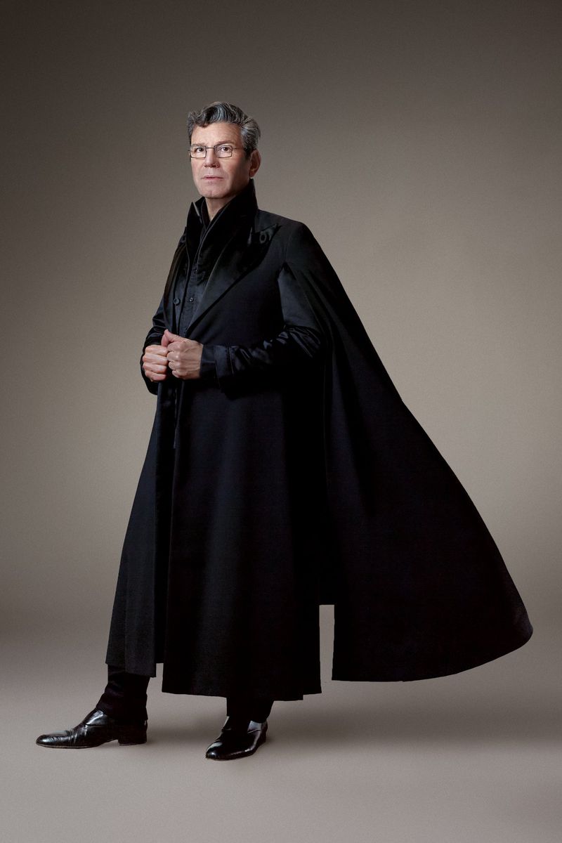 Portrait of a mature adult male wearing a black cape coat and leather shoes in a studio with a neutral background.
