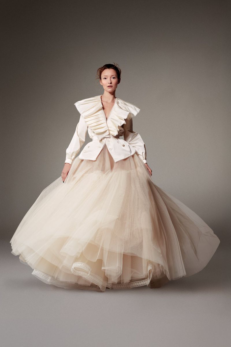 Full-length fashion shot of a woman in a cream-colored, tulle gown with a white ruffled top, posing in a studio setting with soft lighting.