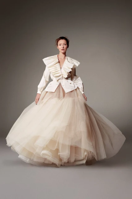 Full-length fashion shot of a woman in a cream-colored, tulle gown with a white ruffled top, posing in a studio setting with soft lighting.