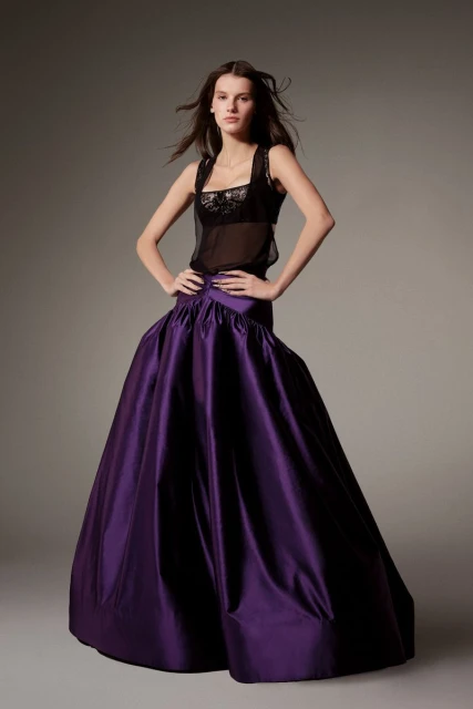 Woman in fashion pose wearing a sheer top and a large, high-waisted purple satin skirt in a studio setting with soft lighting.