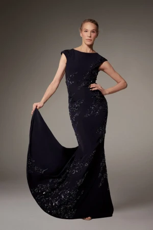Model wearing a dark evening gown with beaded embroidery, posing against a neutral grey studio backdrop.