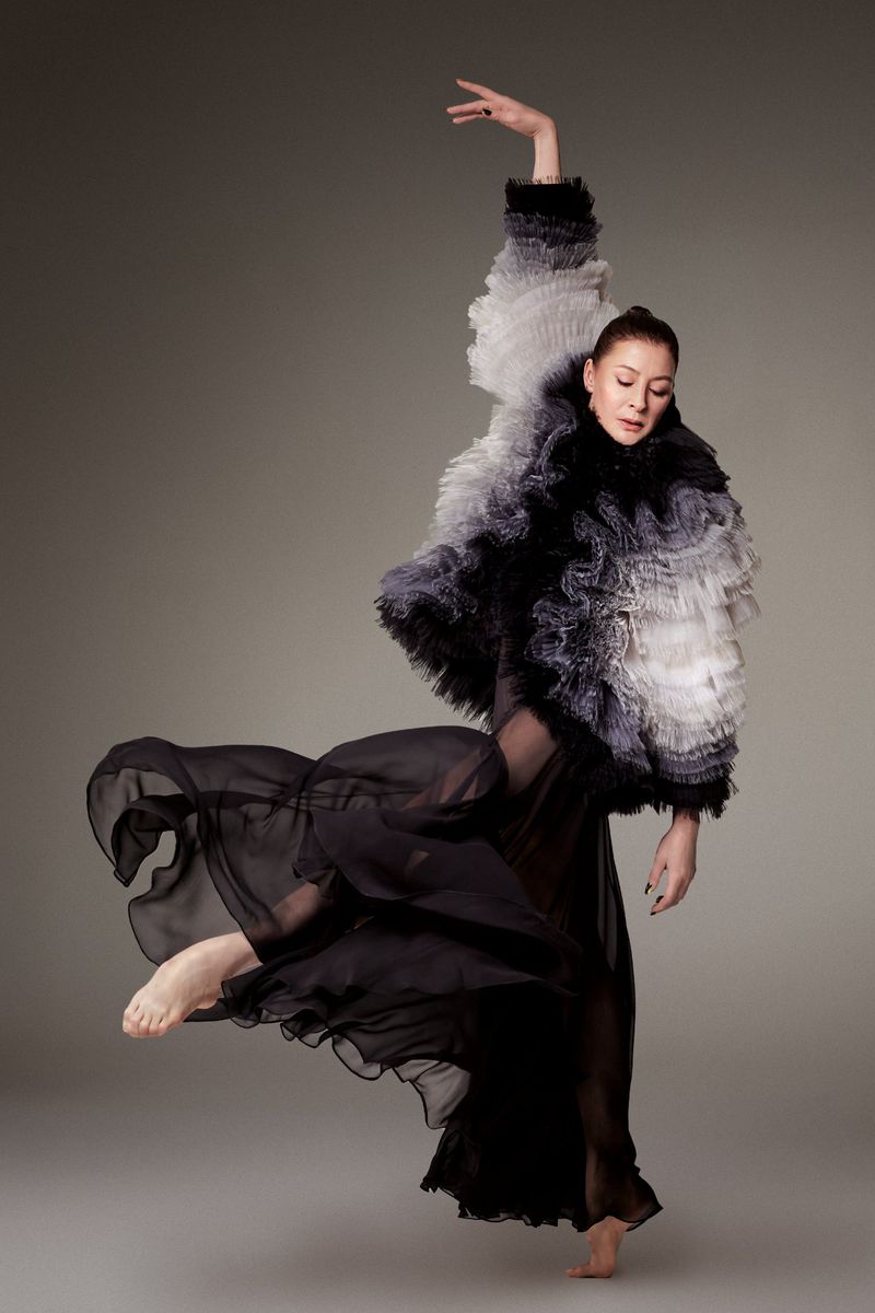 Woman in flowing black dress and ruffled ombre jacket strikes dynamic pose in studio. The image showcases fashion, movement, and grace.