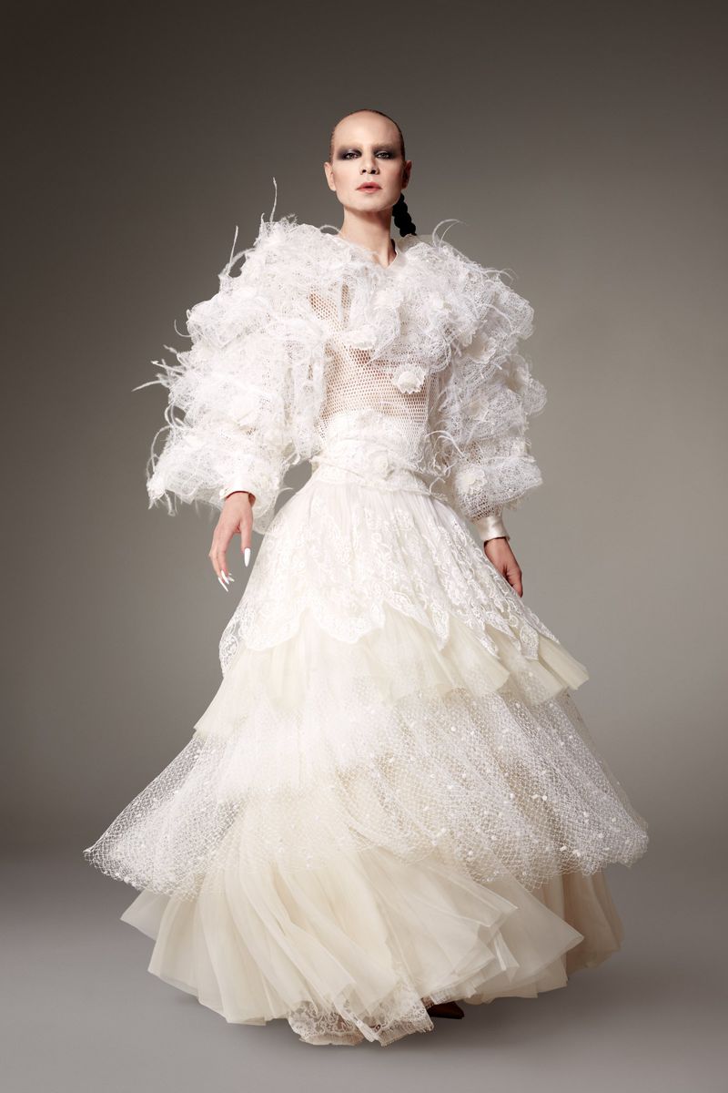 Full-length studio shot of an adult female fashion model wearing an elaborate tiered white gown with feather-like accents and sleek hairstyle.