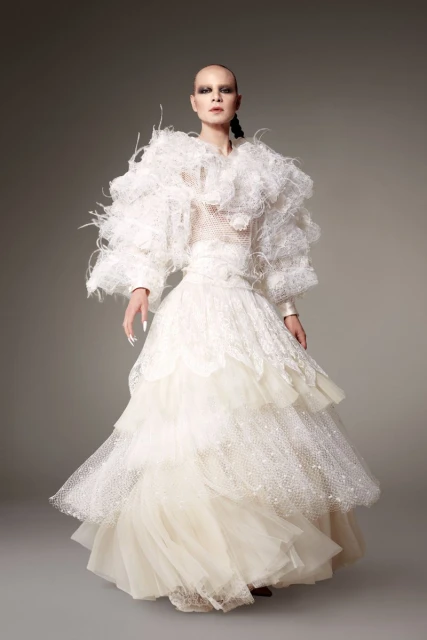 Full-length studio shot of an adult female fashion model wearing an elaborate tiered white gown with feather-like accents and sleek hairstyle.