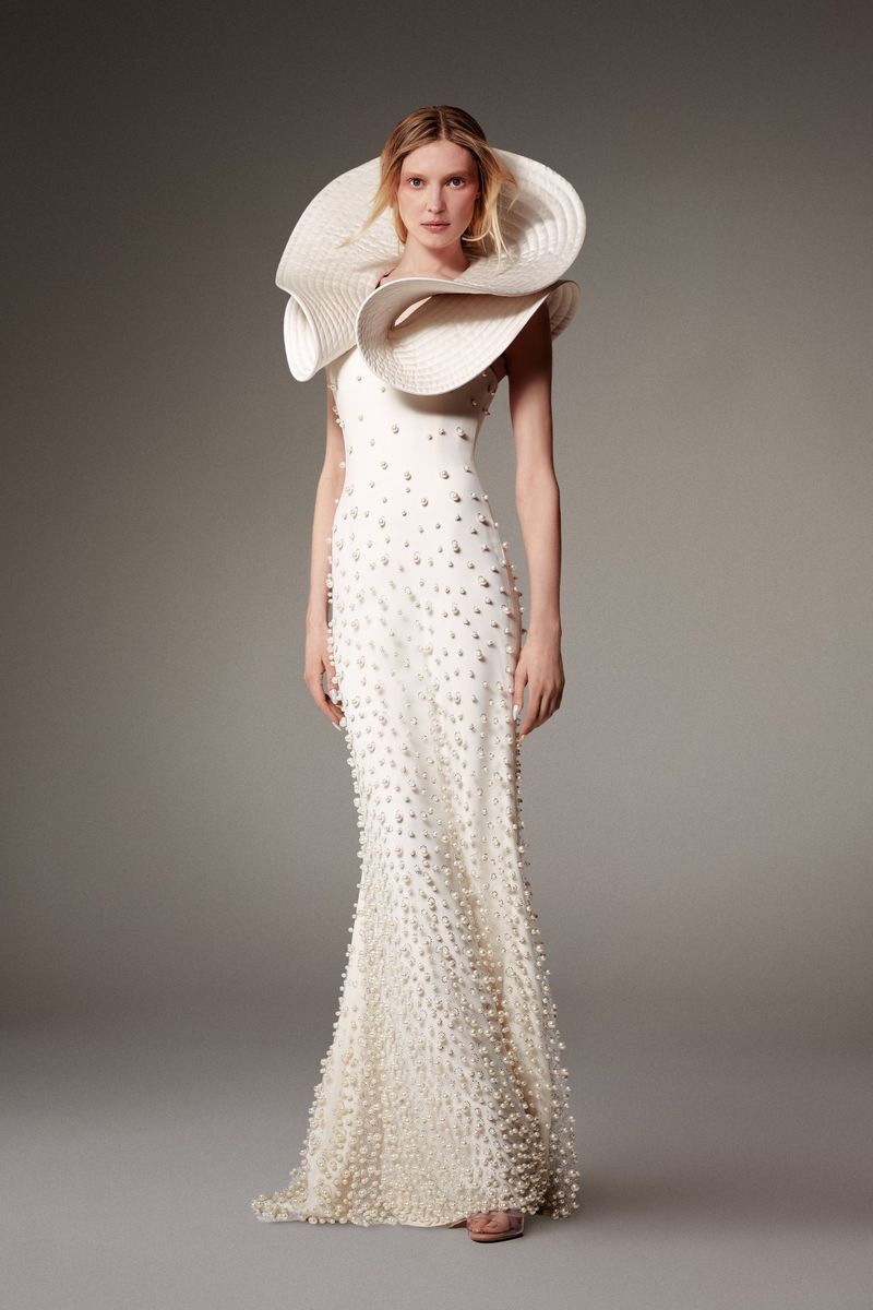 Elegant woman in a white gown with pearl embellishments and a large, textured hat posing in a photo studio with a neutral background.