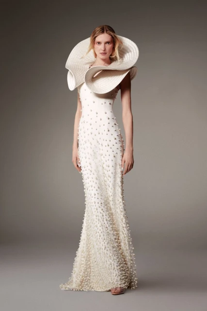Elegant woman in a white gown with pearl embellishments and a large, textured hat posing in a photo studio with a neutral background.